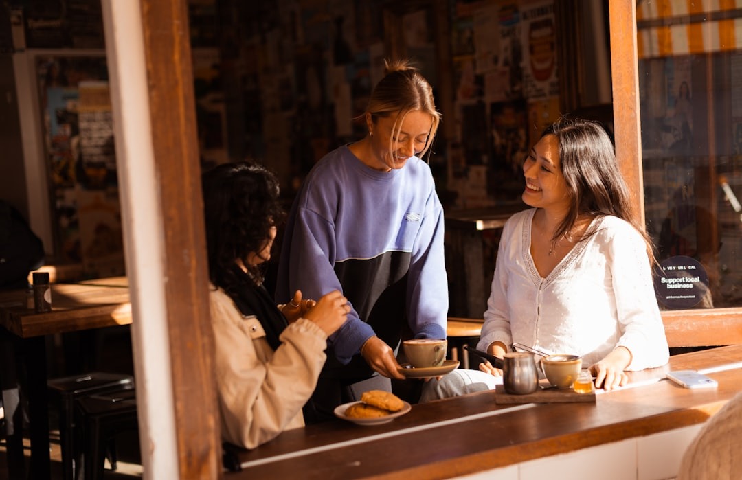 Three smiling women interact at a cafe counter while serving coffee.